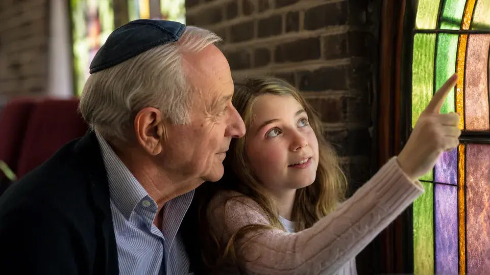 A grandfather and granddaughter looking at stained glass in a synagogue.